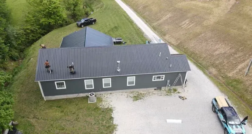 Aerial view of a gray house with a dark roof, gravel driveway, and a parked pickup beside fields