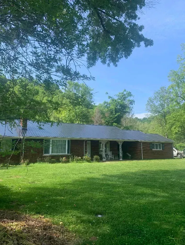 Single-story brick house with a gray roof, partly shaded by trees, overlooking a green lawn under a blue sky