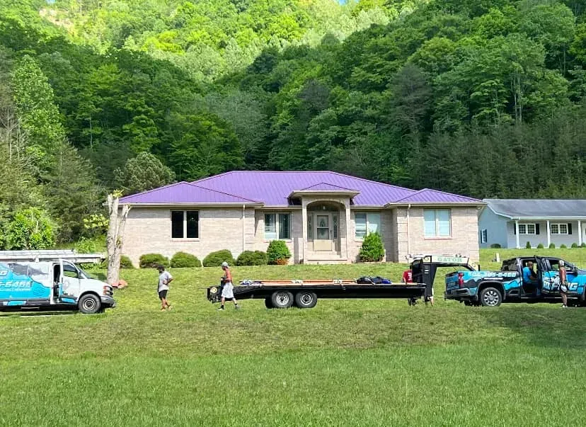 Workers and vehicles outside a small house with a purple roof beside a wooded hillside