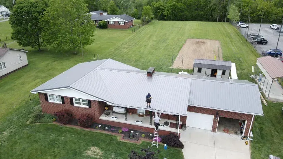 Aerial view of a brick house with white metal roof, front porch, driveway, and large green yard.