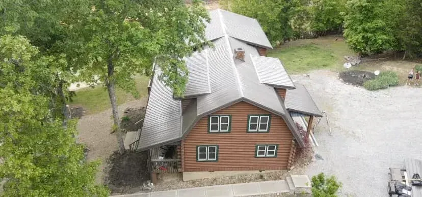Aerial view of a red-brick house with a steep gray roof surrounded by trees and gravel driveway