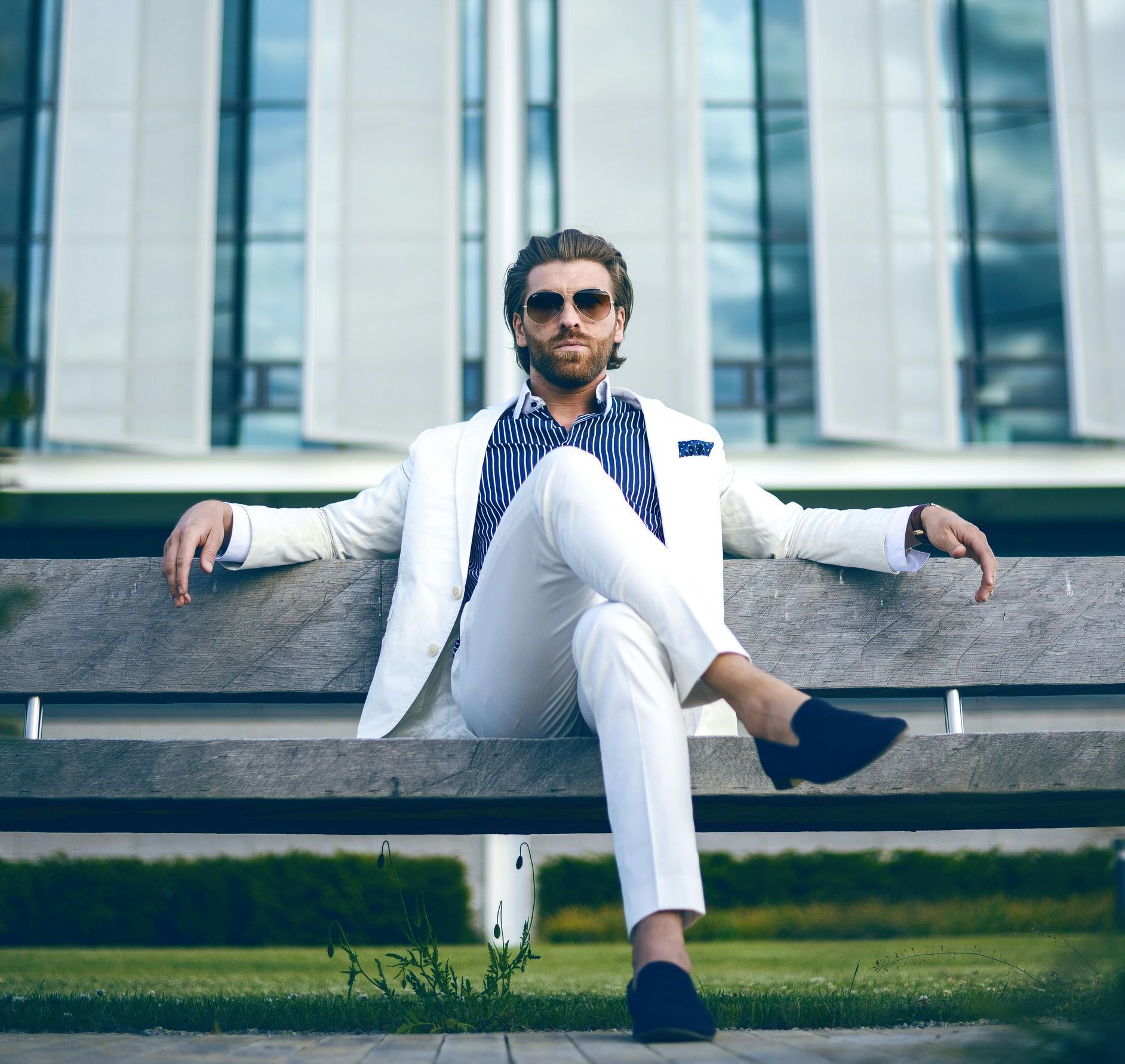 Man in wit pak en zonnebril zit op een bankje voor een gebouw.