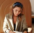 Een schrijvende vrouw, gekleed in een blazer en bandana, in een kamer met bruine muren, kijkt peinzend naar beneden.