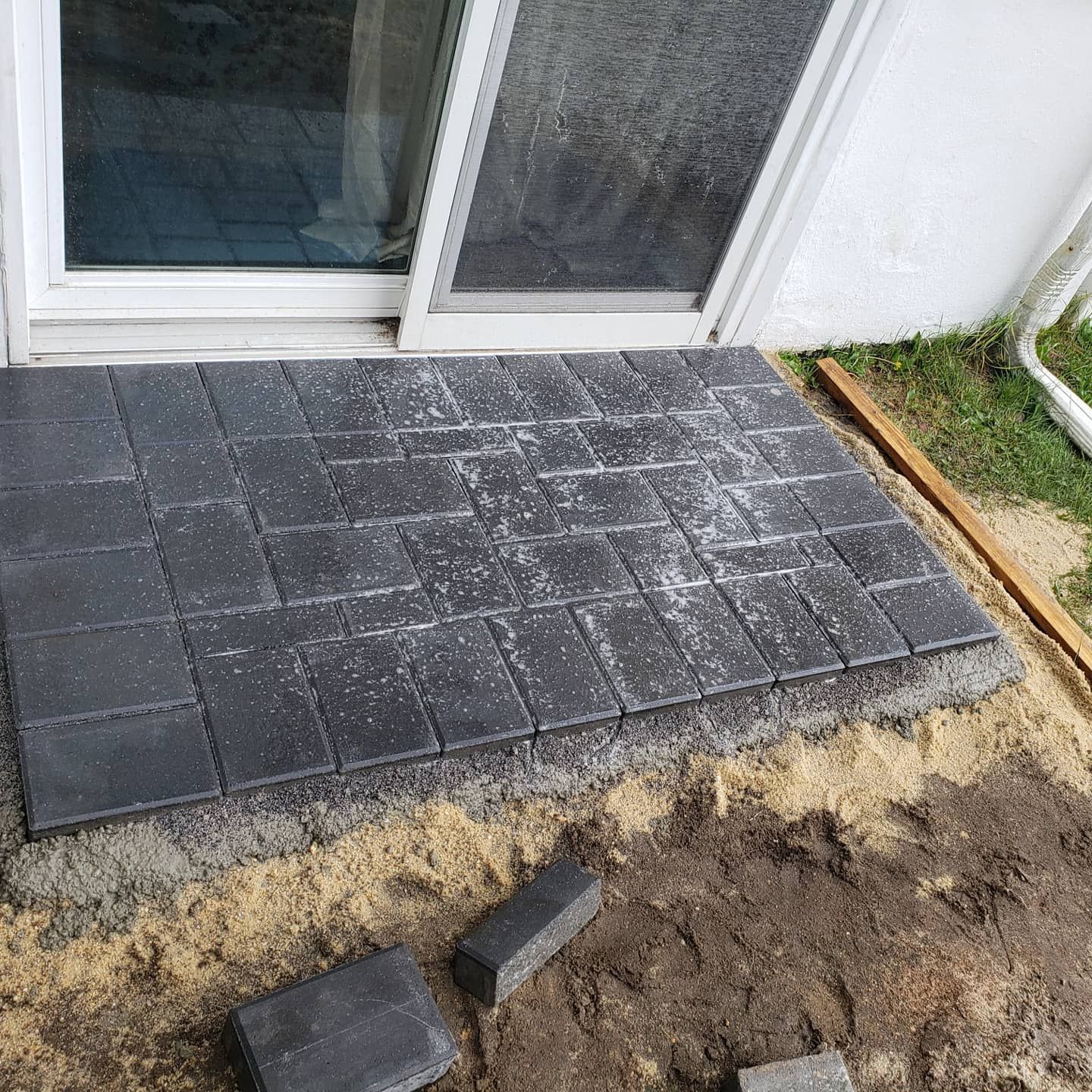 Dark pavers being installed outside a sliding glass door. The pavers are set on sand, and tools are nearby.