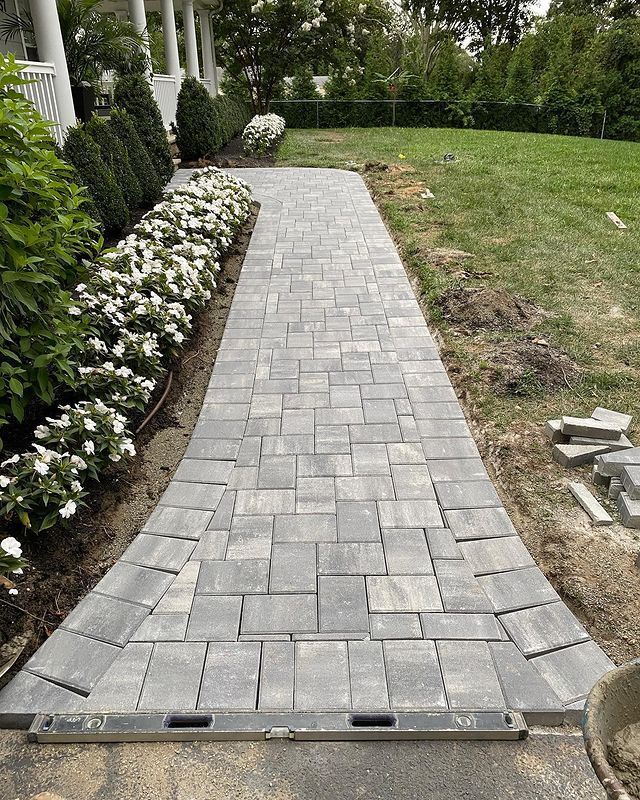 A newly constructed brick pathway leading towards a house, bordered by flowers and grass.