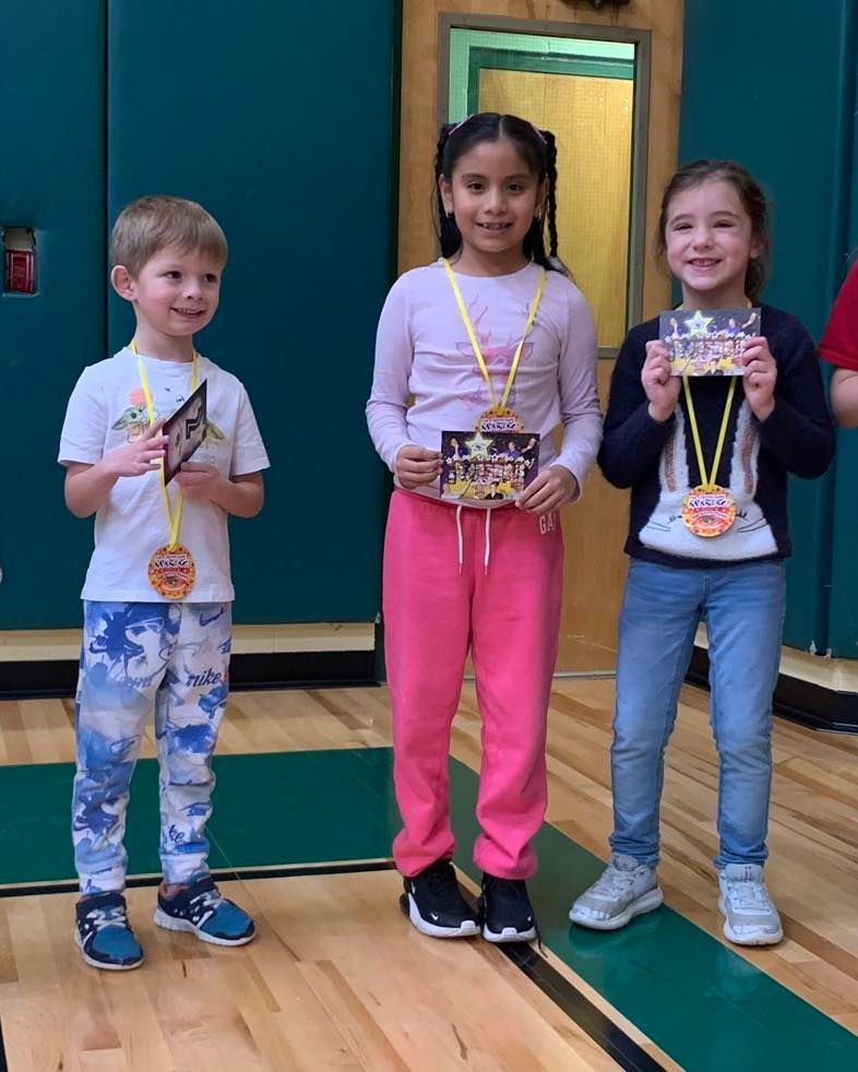 Three children are standing in a gym holding medals
