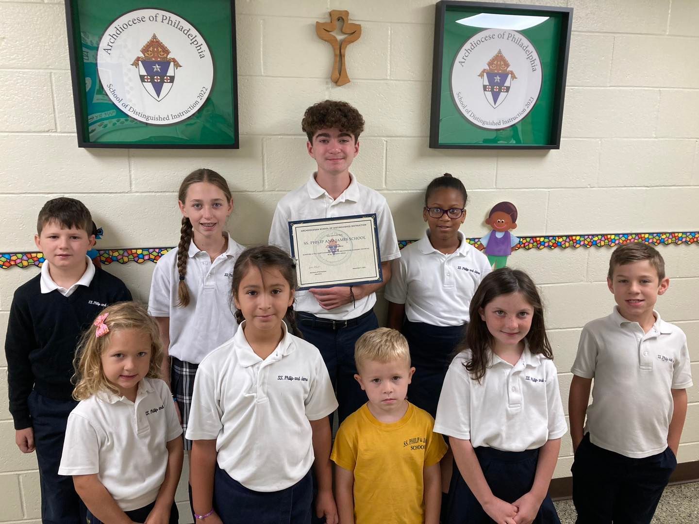 Group of students in uniforms pose for a photo, holding a certificate, in front of school emblems.