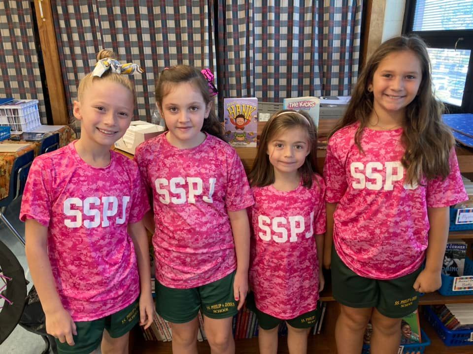 Four young girls wearing pink shirts and green shorts are standing next to each other in a classroom.