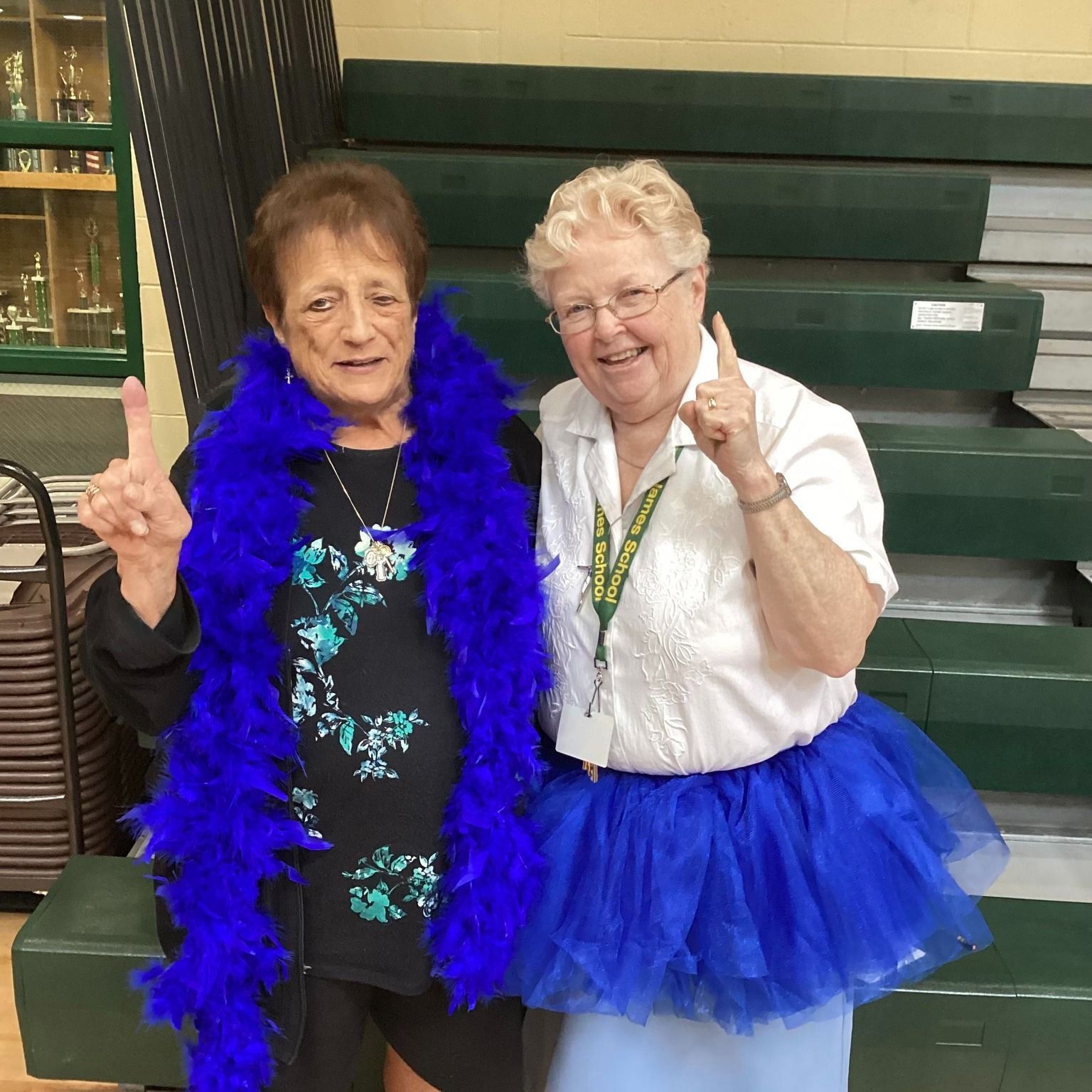 Two women are posing for a picture and one is wearing a blue boa and the other is wearing a blue tutu.