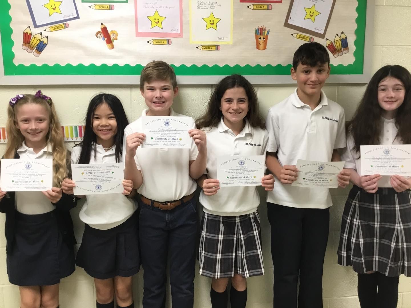 A group of children in school uniforms are holding certificates in front of a bulletin board.