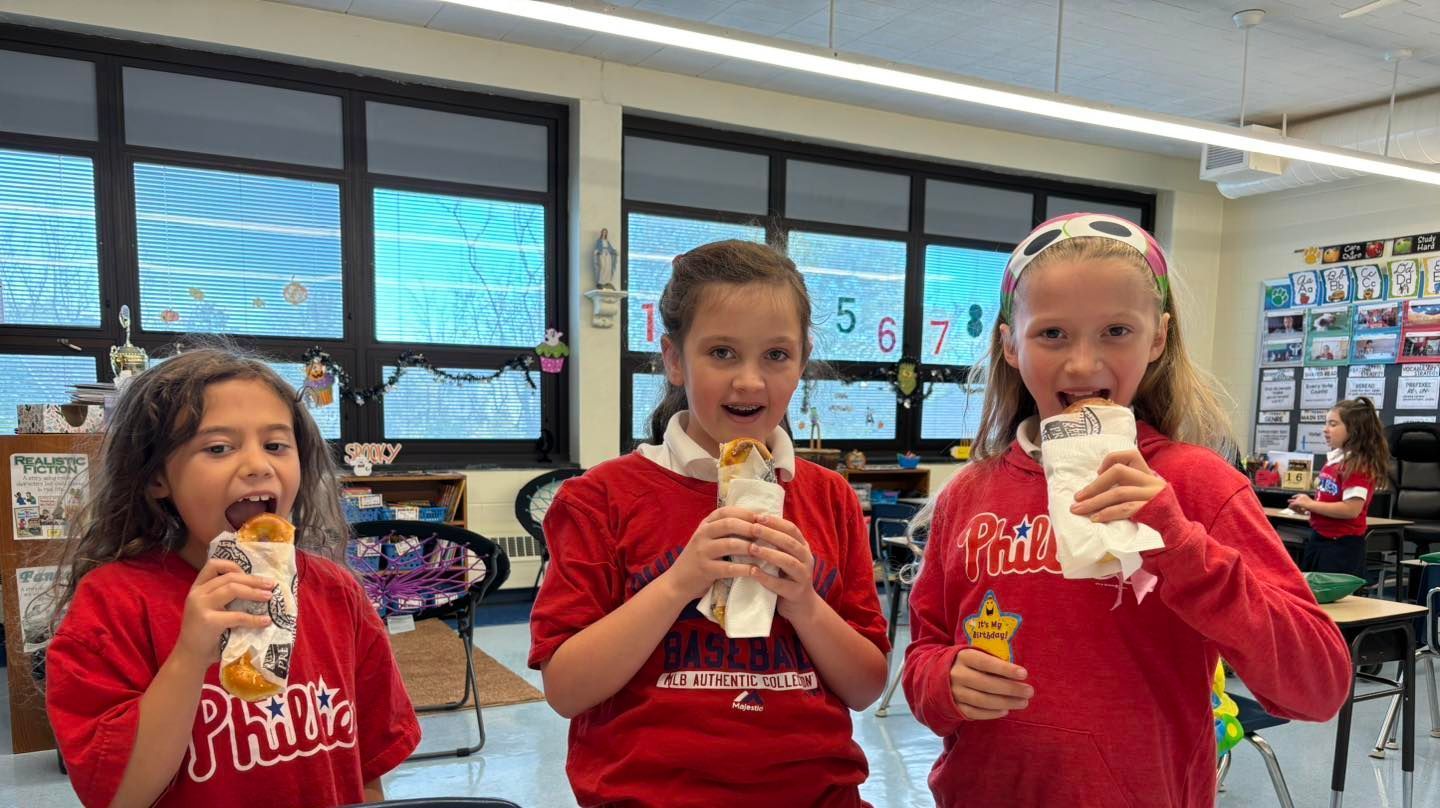 Three young girls in red shirts are eating sandwiches in a classroom.