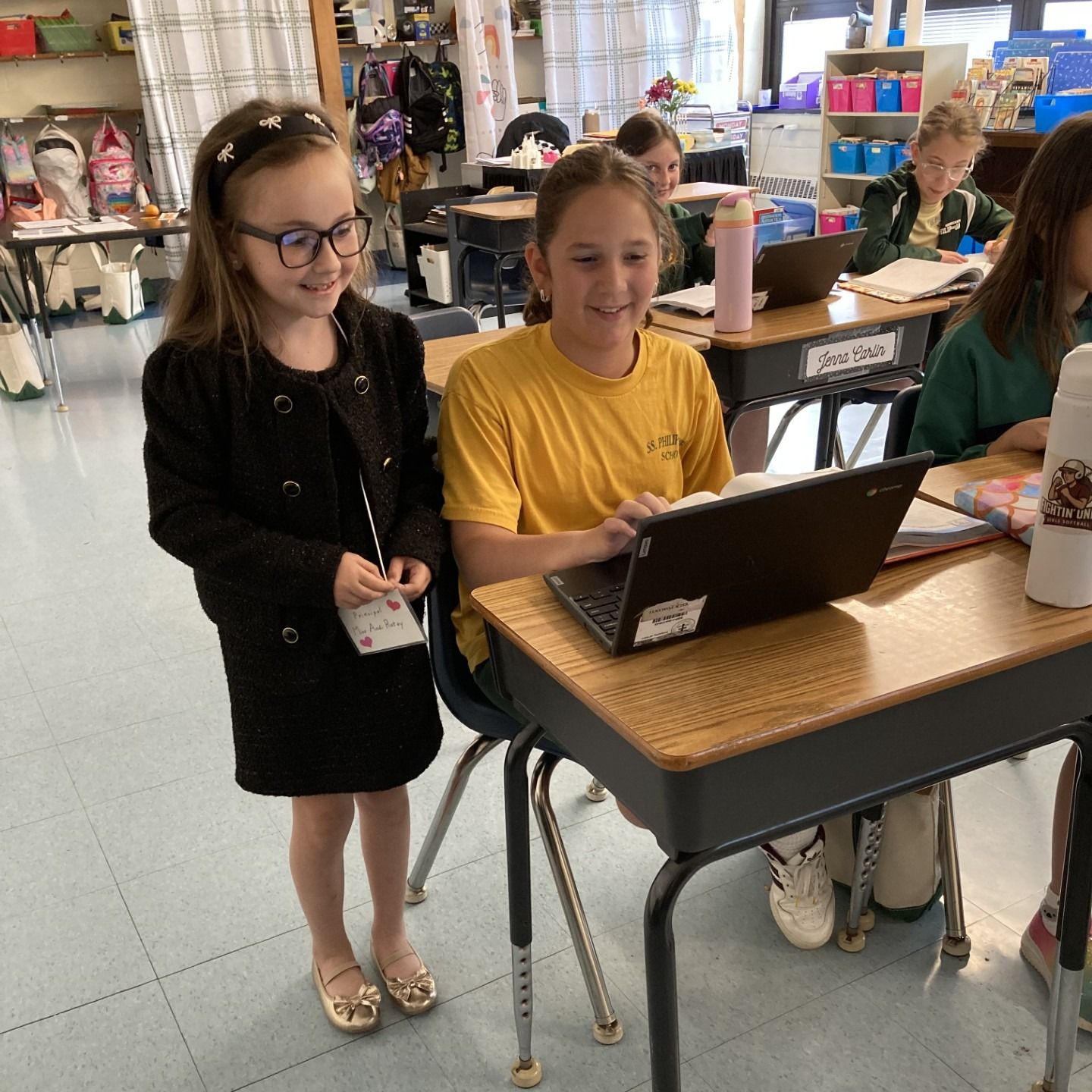 Two young girls in a classroom, one using a laptop, the other smiling beside her.