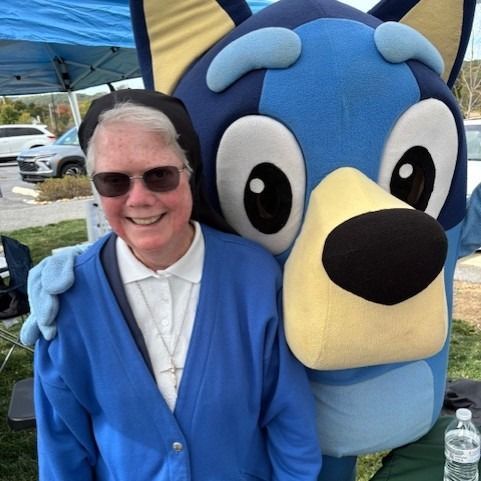 A woman in a blue cardigan and sunglasses smiles next to a Bluey mascot outdoors.