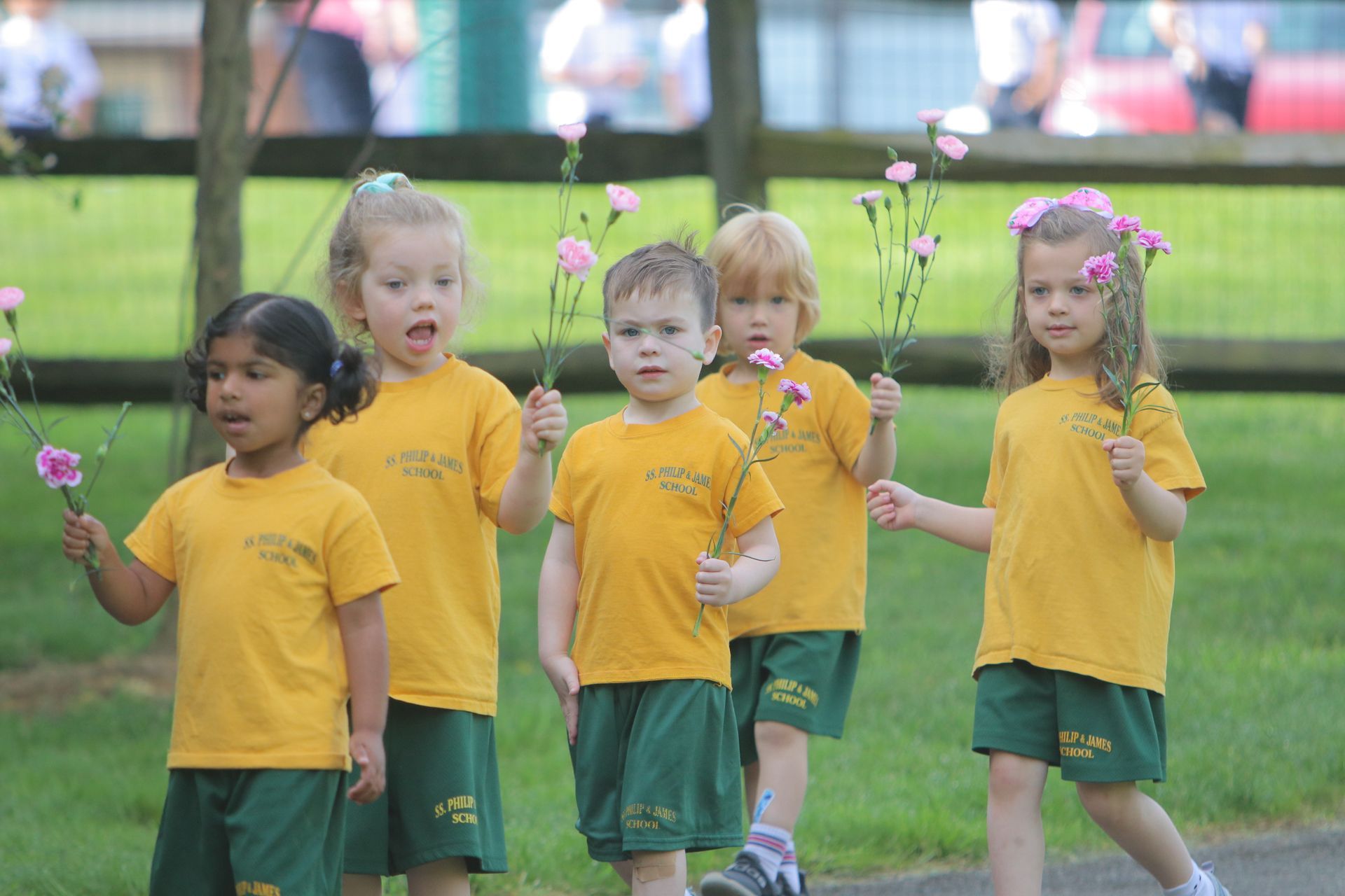 A group of children wearing yellow shirts and green shorts are holding flowers.