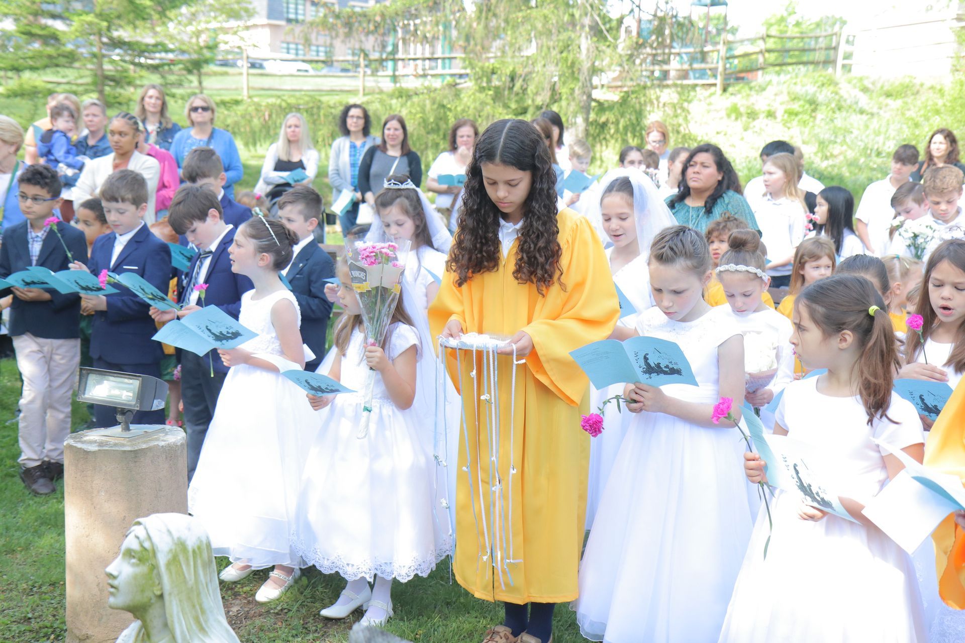 A group of children in white dresses are singing in a park.