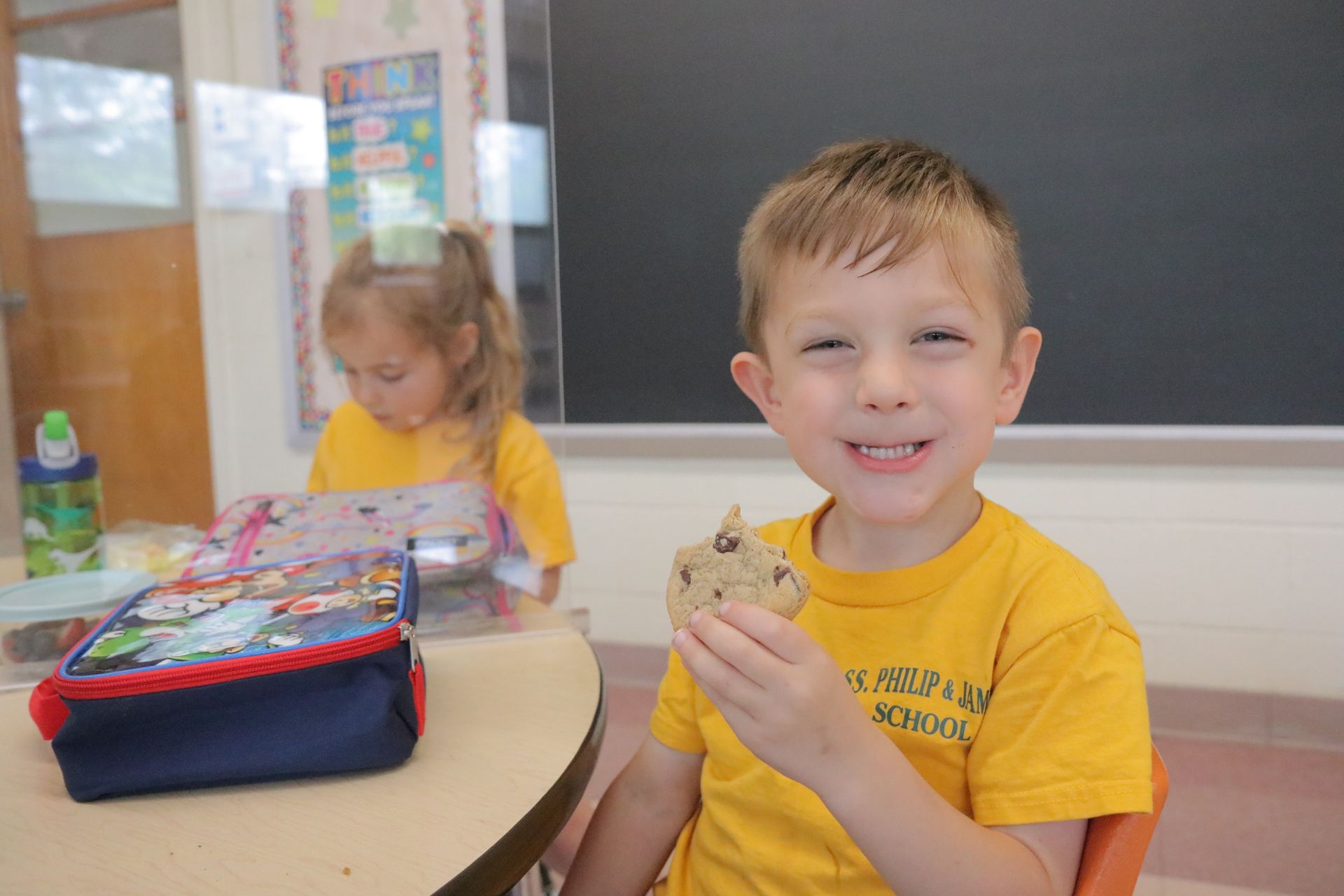 A young boy is sitting at a table eating a cookie in a classroom.