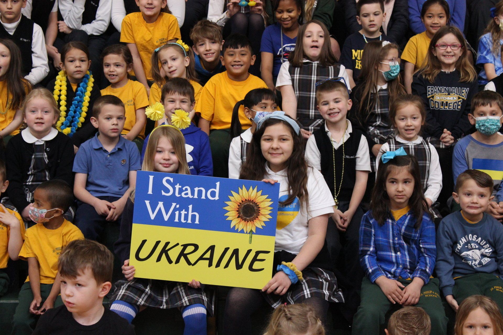 A girl is holding a sign that says `` i stand with ukraine '' in front of a large group of children.