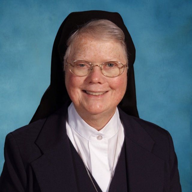 A nun wearing glasses and a black veil smiles for the camera