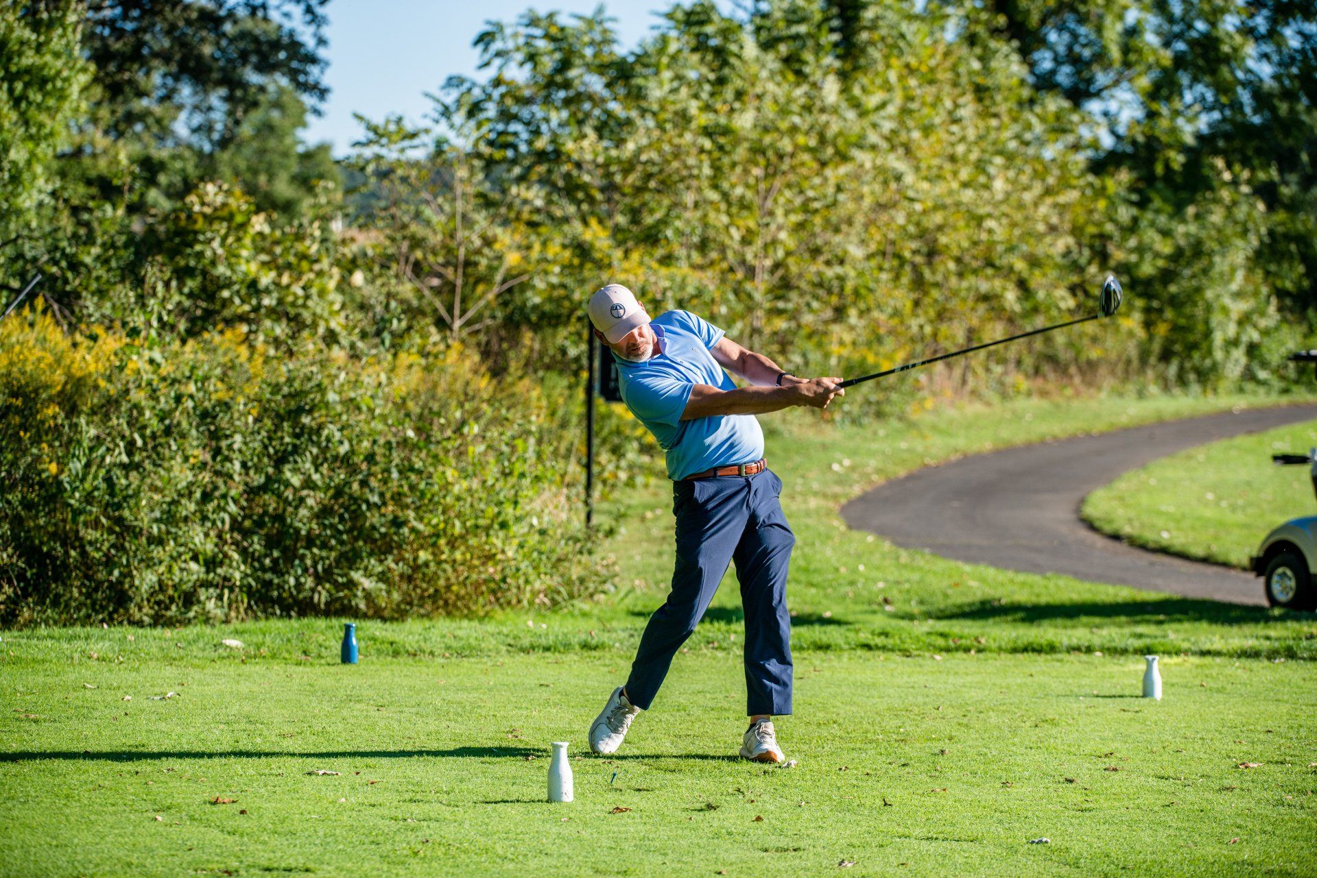 Golfer in blue shirt swings club on green course. A golf cart is parked in the background.