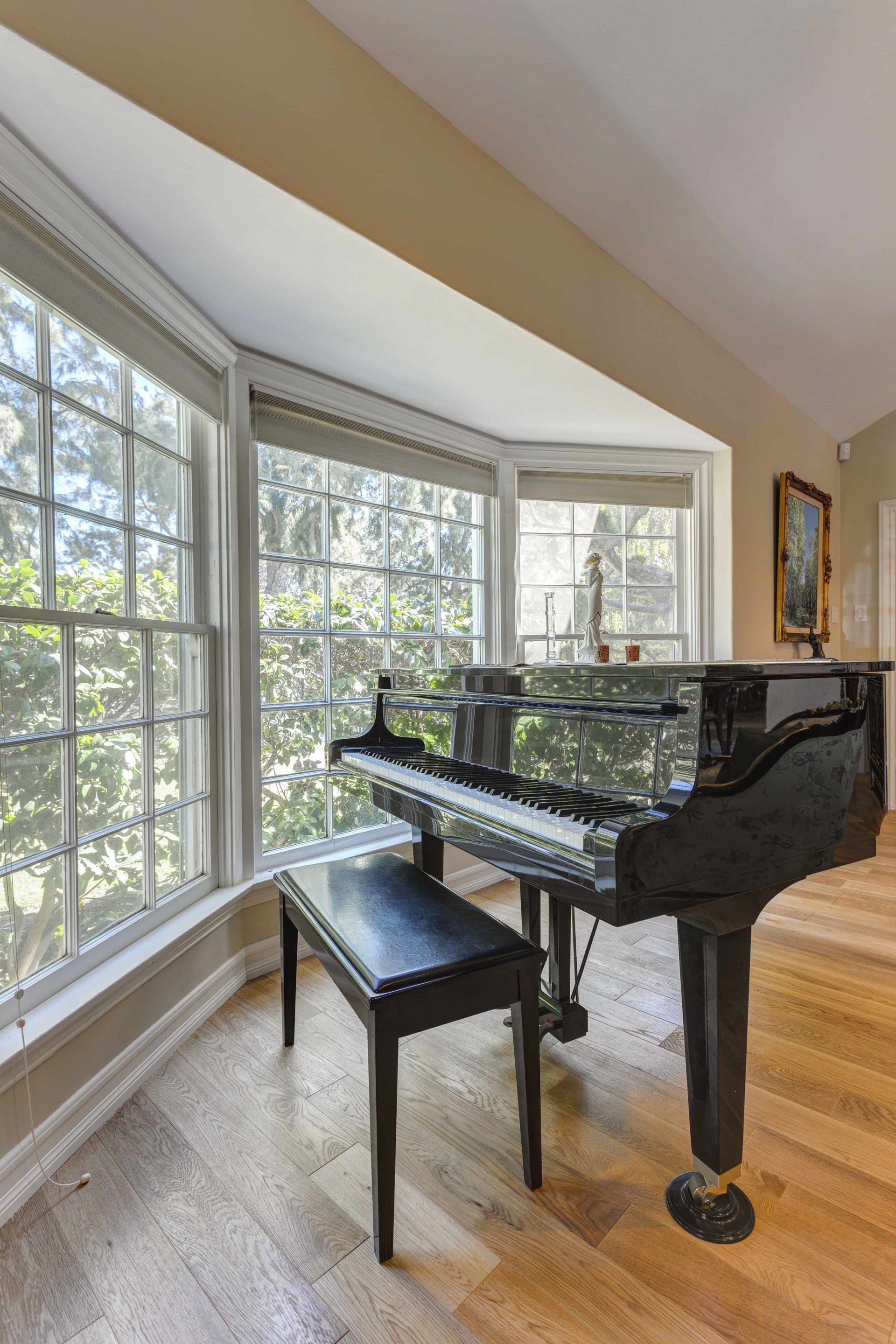 Black grand piano by large bay windows in a bright room with wood floors.