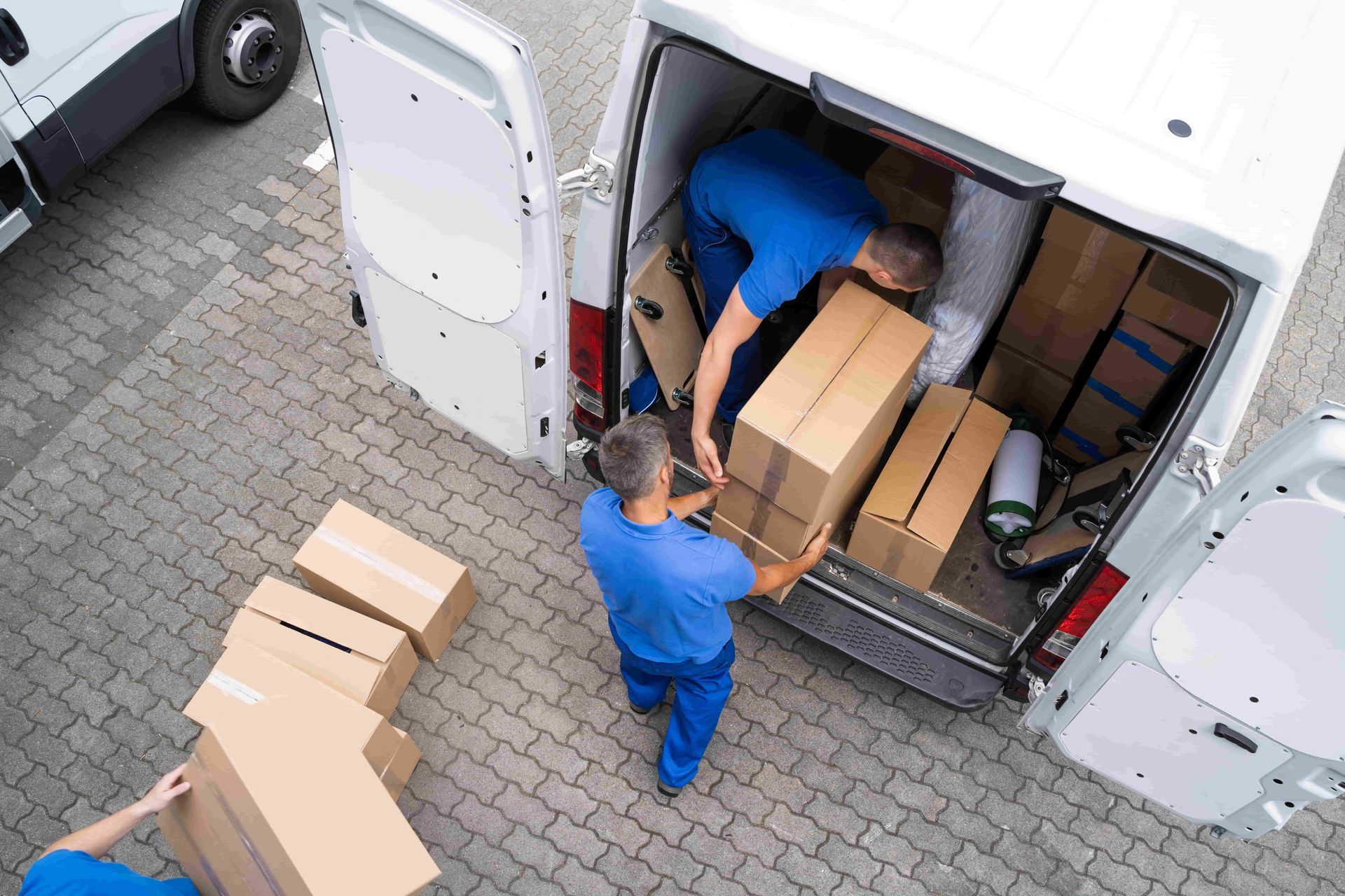 Two workers loading cardboard boxes into a delivery van from a paved lot.
