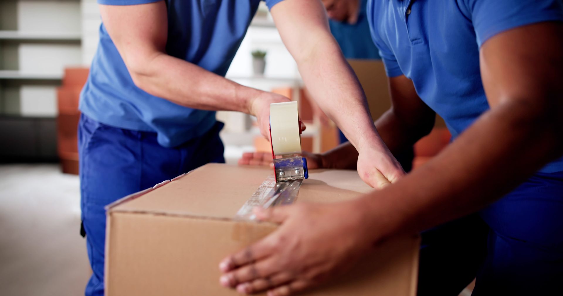 Two people in blue uniforms taping a cardboard shipping box.