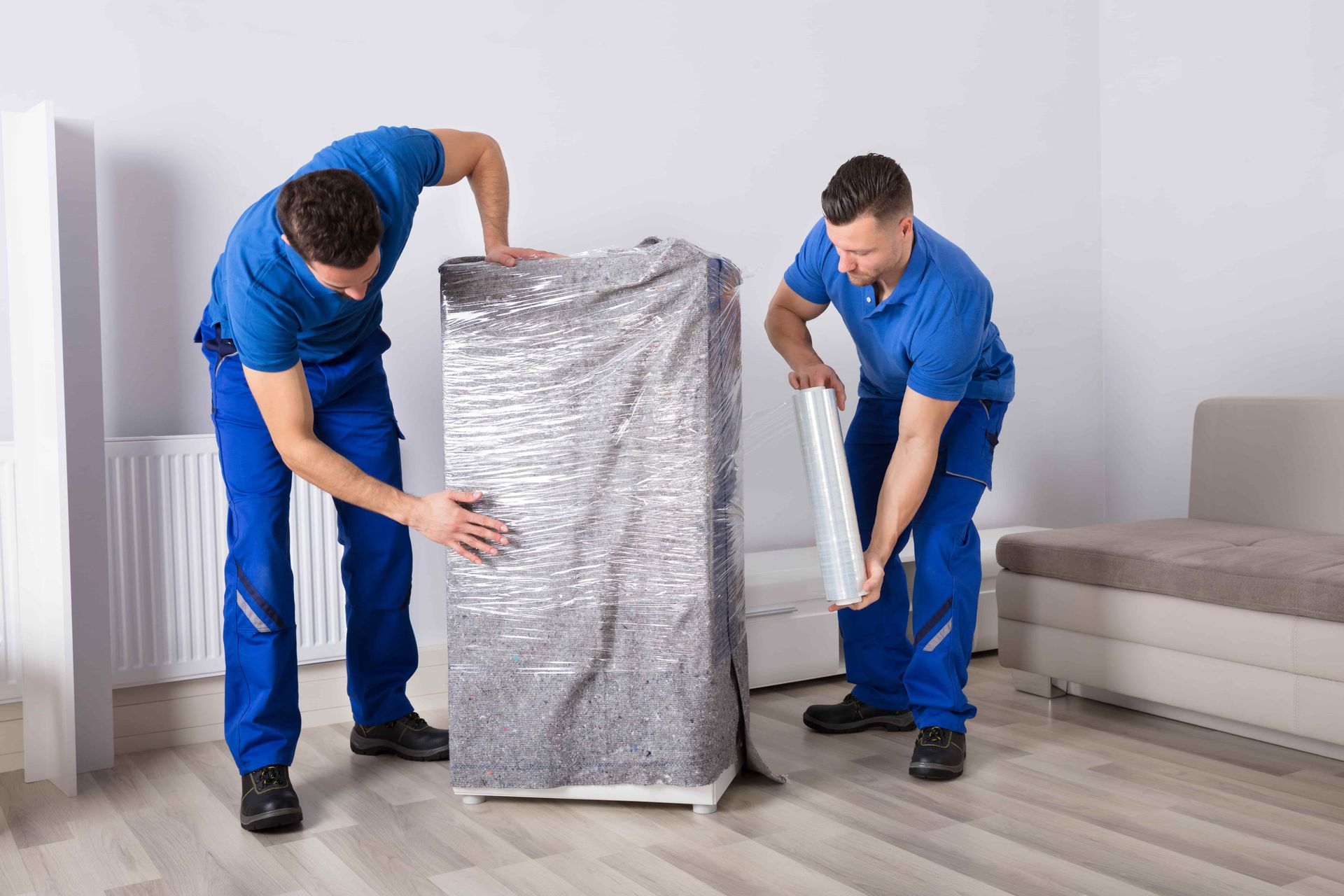 Two movers in blue uniforms wrap a tall object in metallic protective film inside a bright, minimalist room.