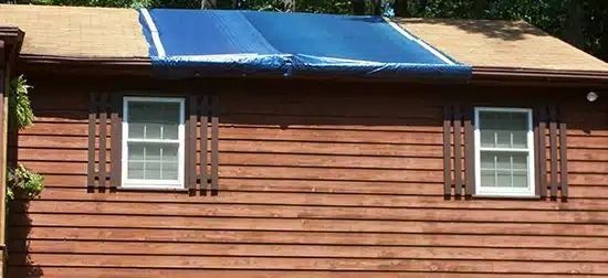 Brown wooden house wall with two small windows and a blue tarp covering part of the roof