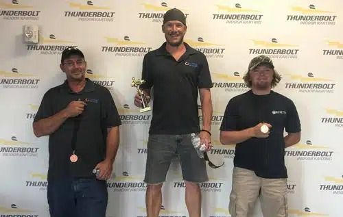 Three men pose holding medals in front of a THUNDERBOLT backdrop, with the center man standing on a podium.