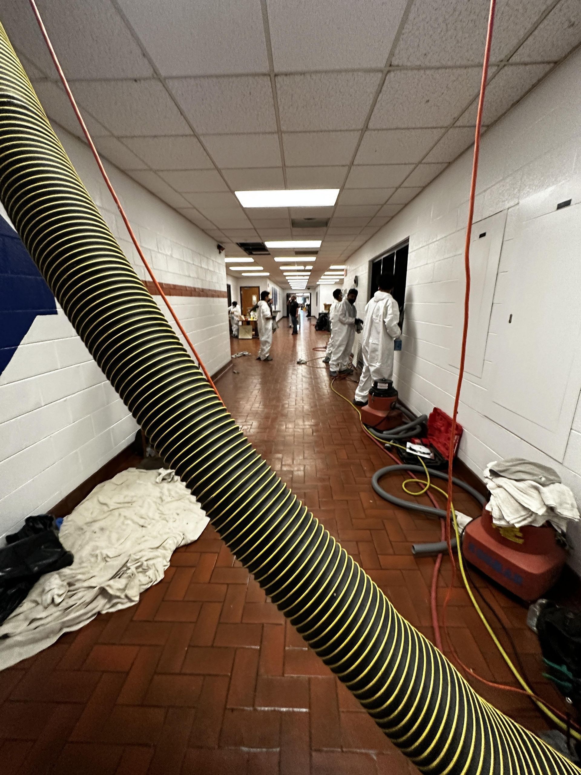 Flooded hallway with workers in white protective suits, hoses, and cleanup equipment on the floor