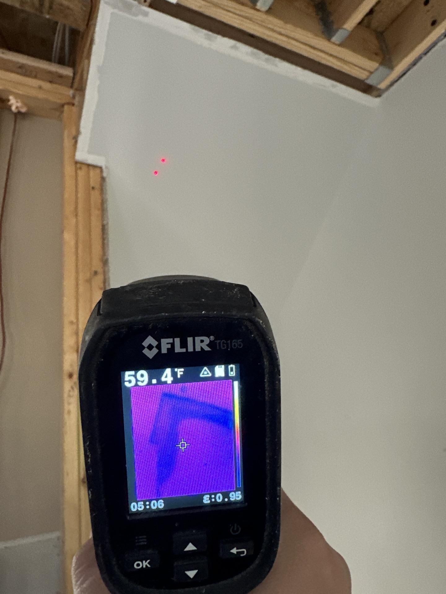FLIR thermal camera aimed at a ceiling corner with exposed wooden framing and a red laser dot