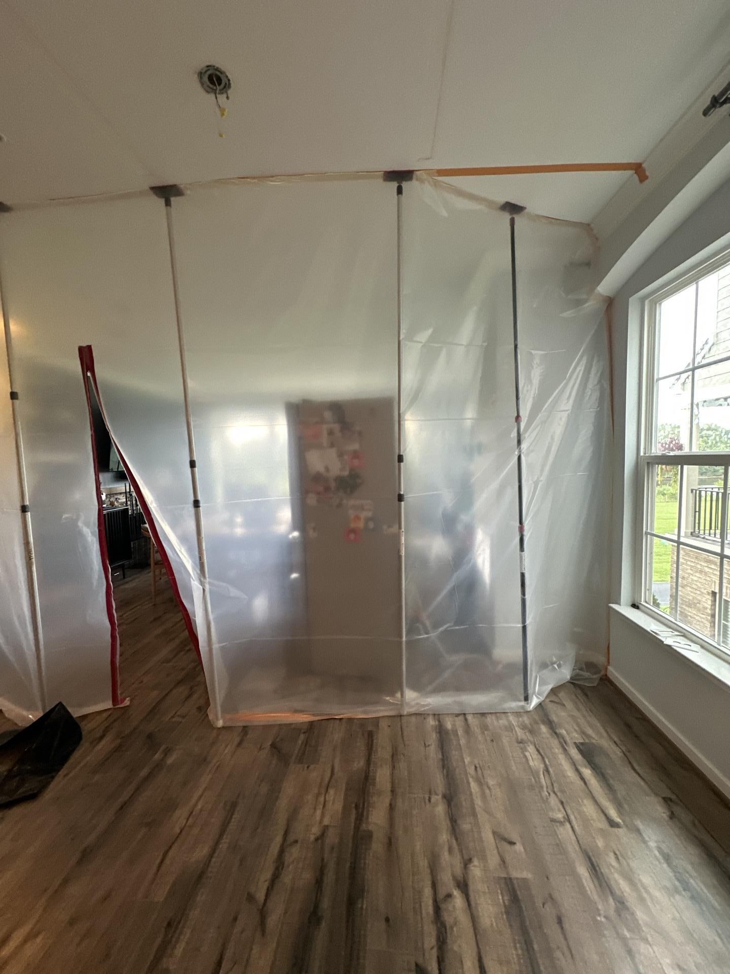 Plastic-sheeted room under renovation with exposed ceiling and wood floor, lit by a window on the right