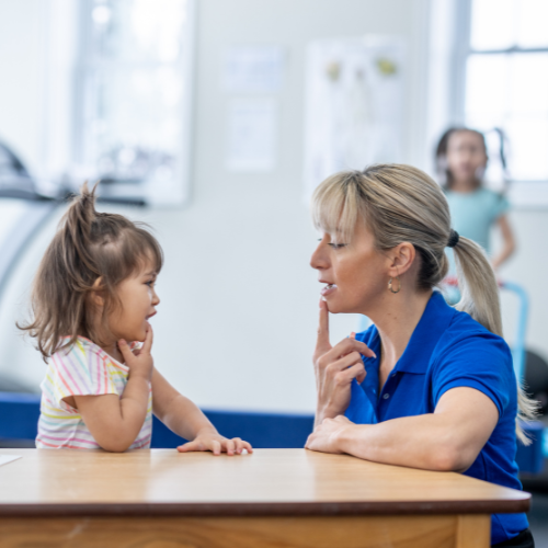 Child in blue shirt with open mouth at a table, looking towards a person out of frame.