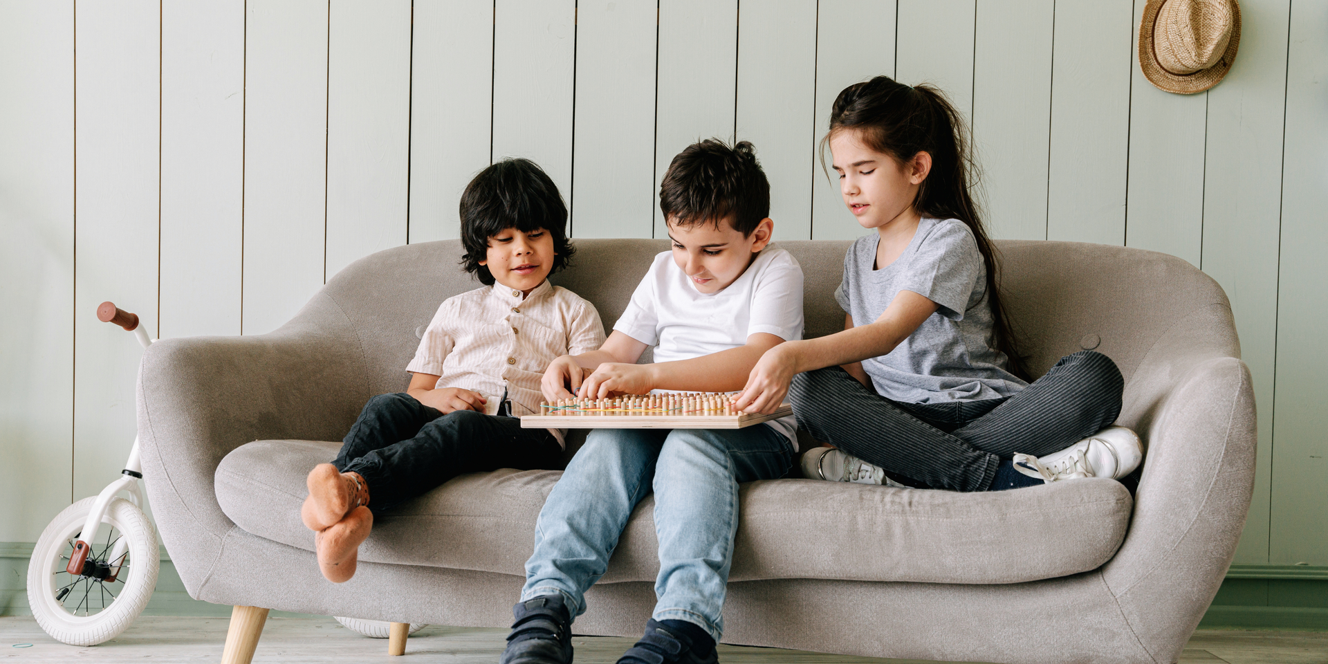 Three children sit on a couch, playing a game with wooden pieces. The room has light green walls.