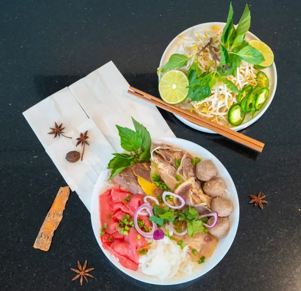Two bowls of colorful noodle soup on a dark table, garnished with lime, herbs, and chopsticks.