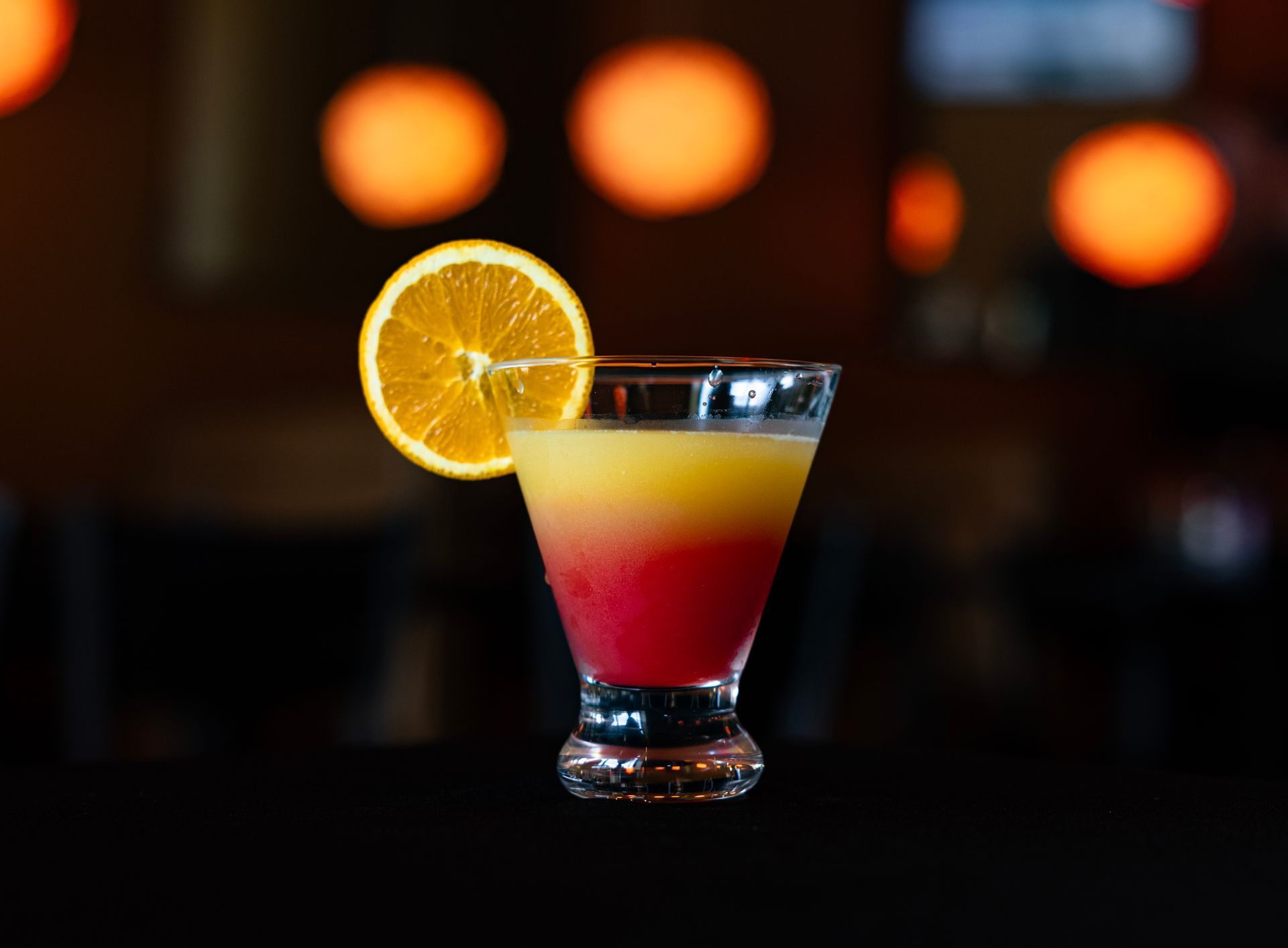 Layered red, yellow, and white cocktail with an orange slice garnish on a dark bar background