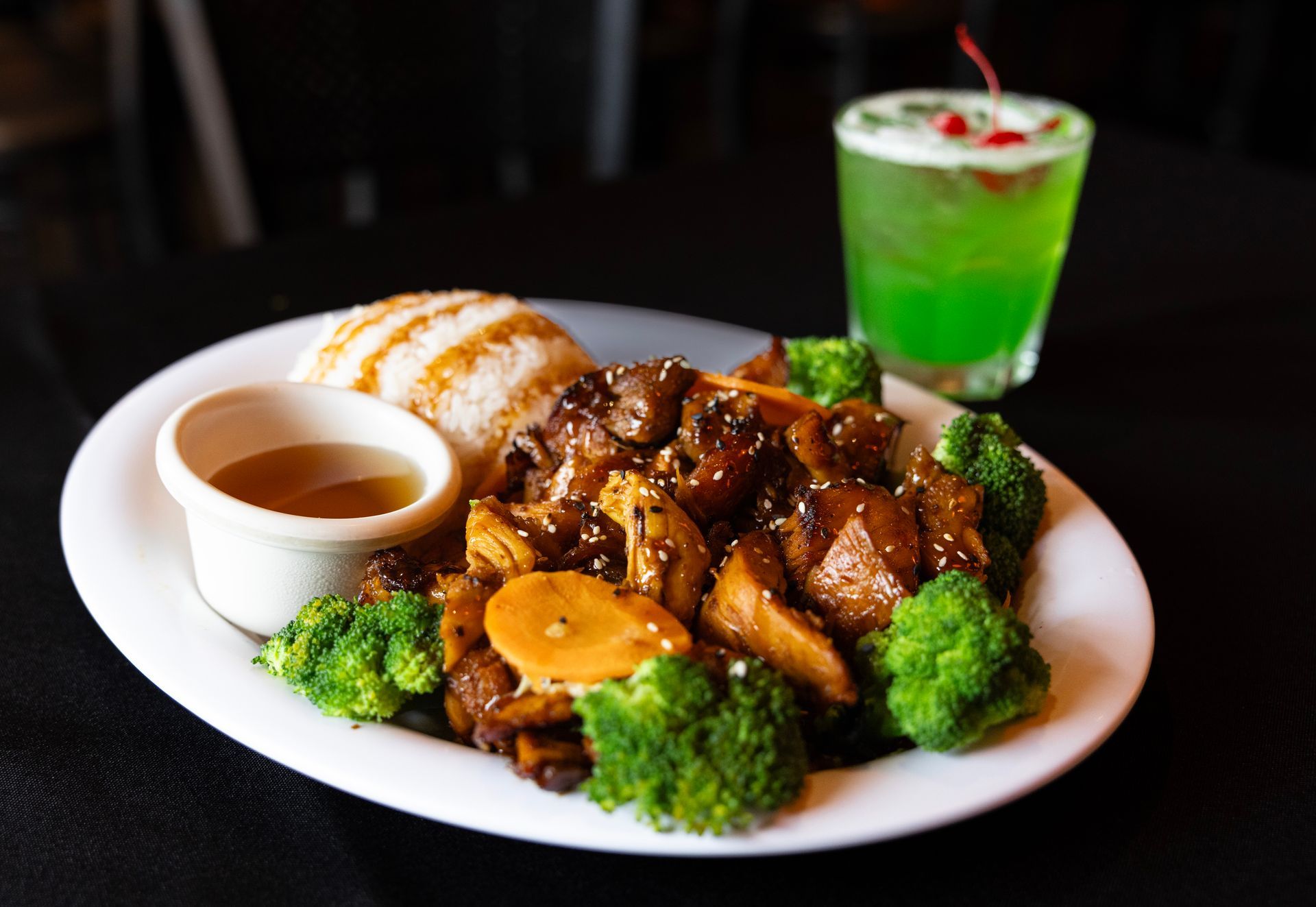 Plated teriyaki chicken with rice, broccoli, and sauce, plus a green drink on a black background