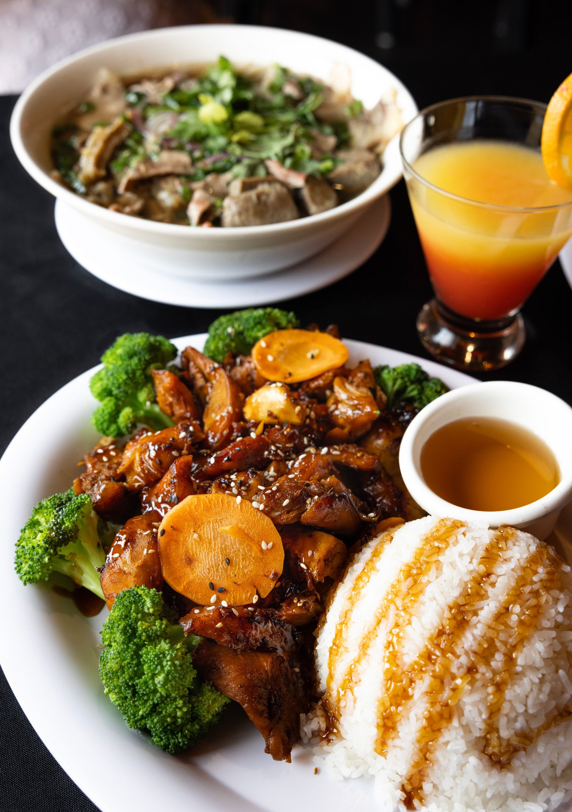 Plated beef with rice and broccoli, bowl of soup, and a glass of orange drink on a table