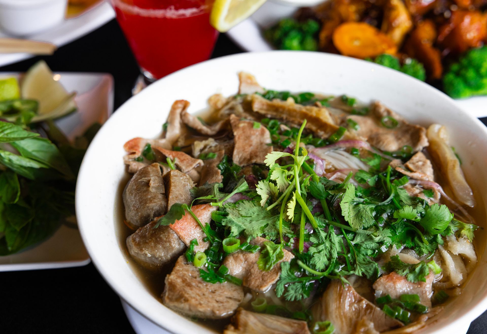 Bowl of beef noodle soup topped with herbs, with a salad and red drink beside it