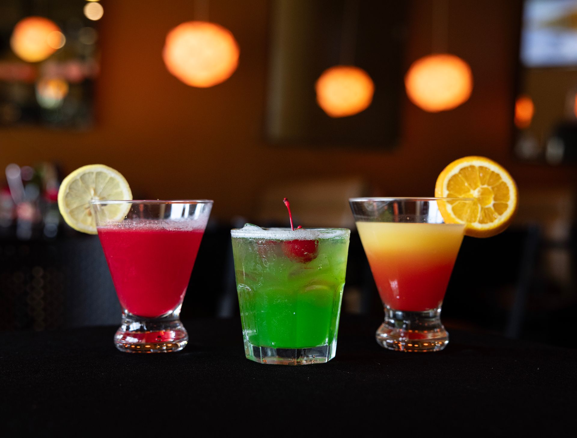 Three colorful cocktails with citrus garnish on a bar, against a softly lit bokeh background