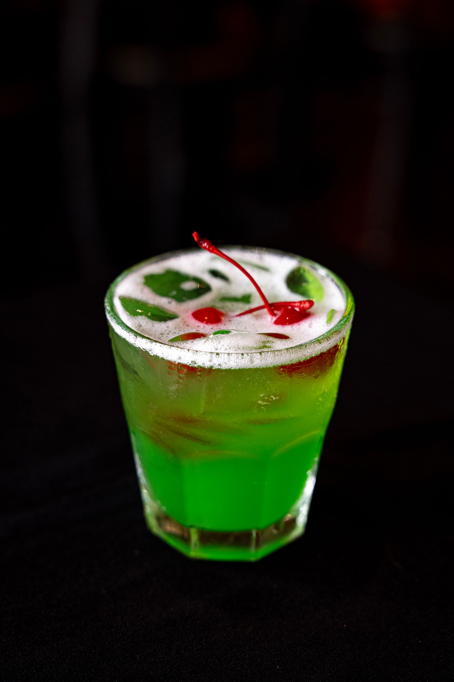 Green cocktail in a short glass with ice and red garnish on a dark background