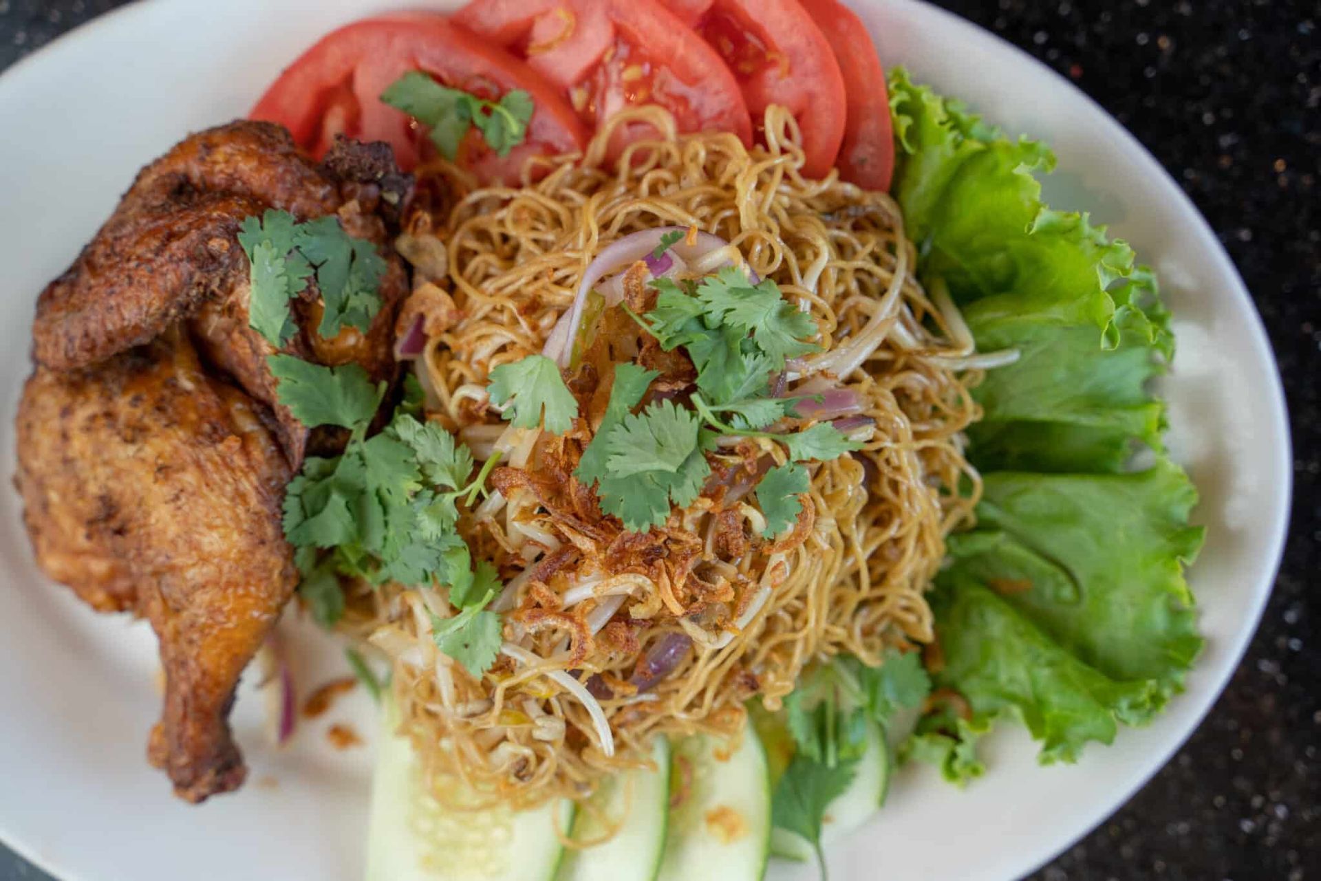 Plate of noodles with fried chicken, sliced tomatoes, lettuce, cucumber, and cilantro garnish