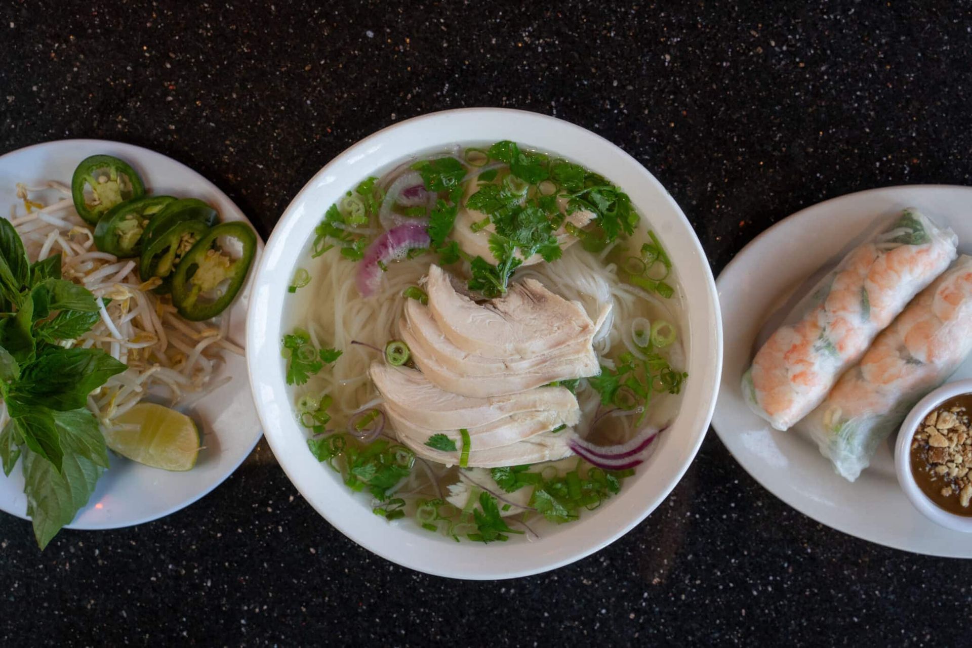 Three plates of Vietnamese food: pho soup, fresh spring rolls, and lime wedges on a dark table