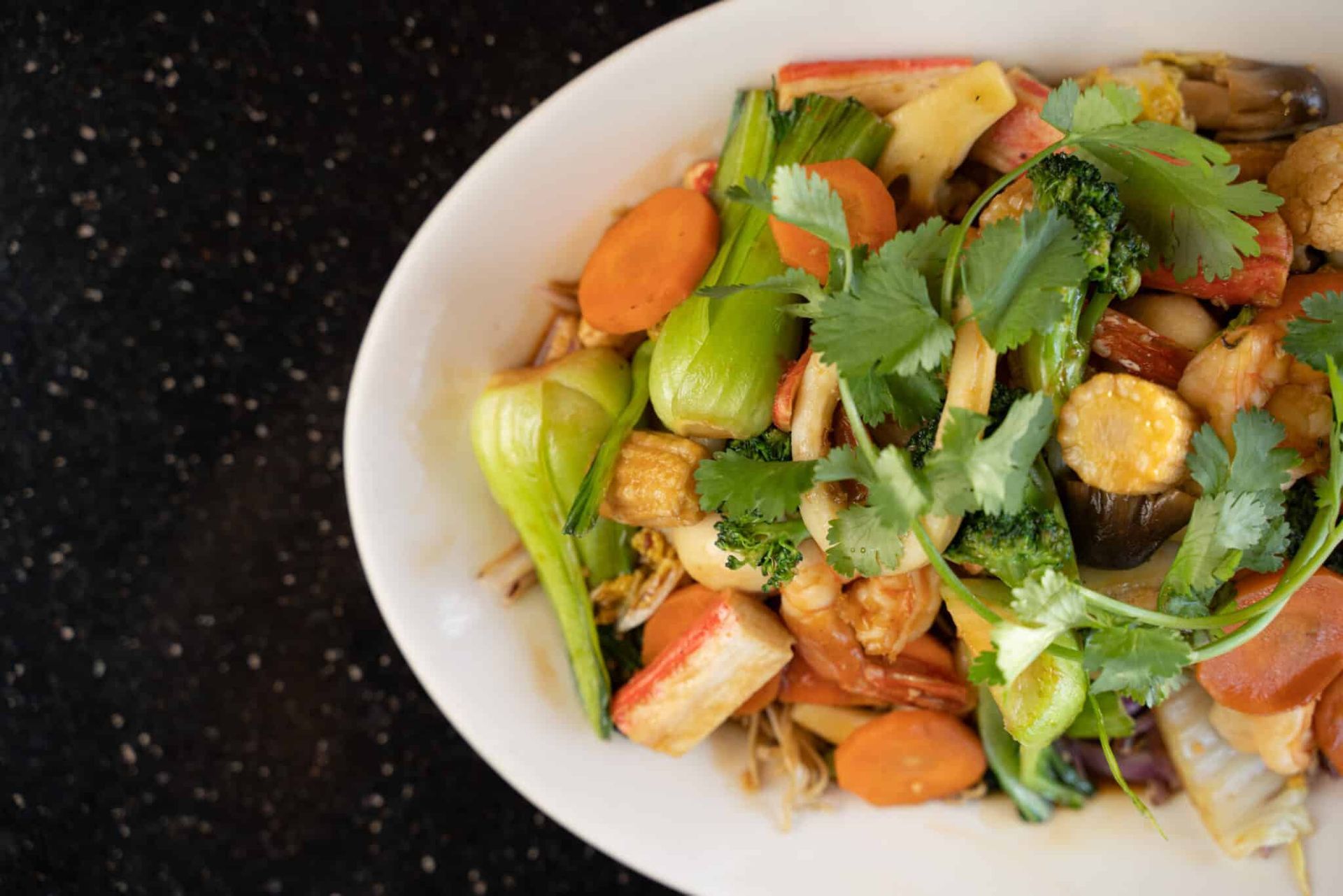 Vegetable noodle salad in a white bowl, topped with cilantro and carrot slices on a dark table