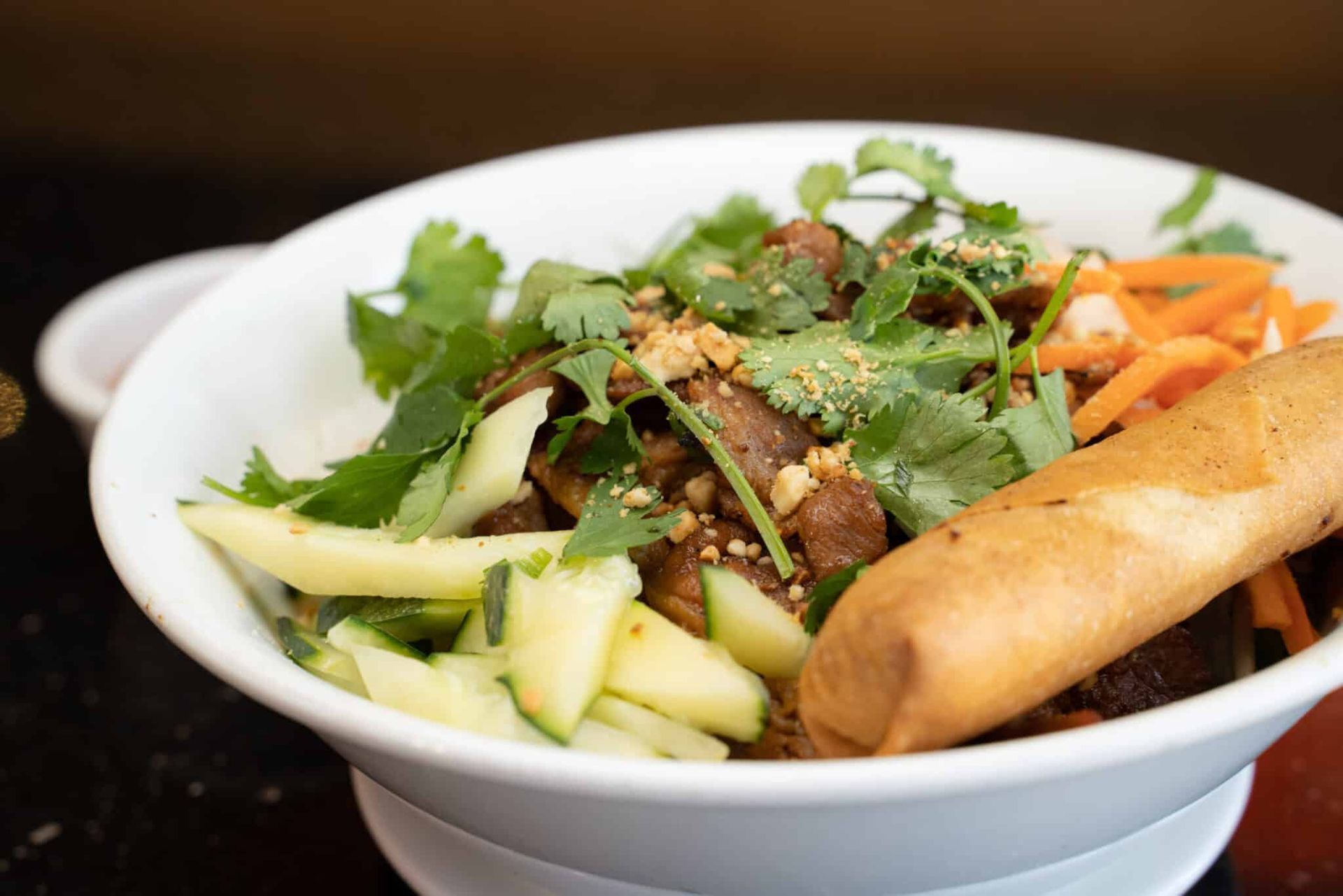 White bowl of fresh Vietnamese vermicelli with herbs, sliced vegetables, grilled meat, and a spring roll