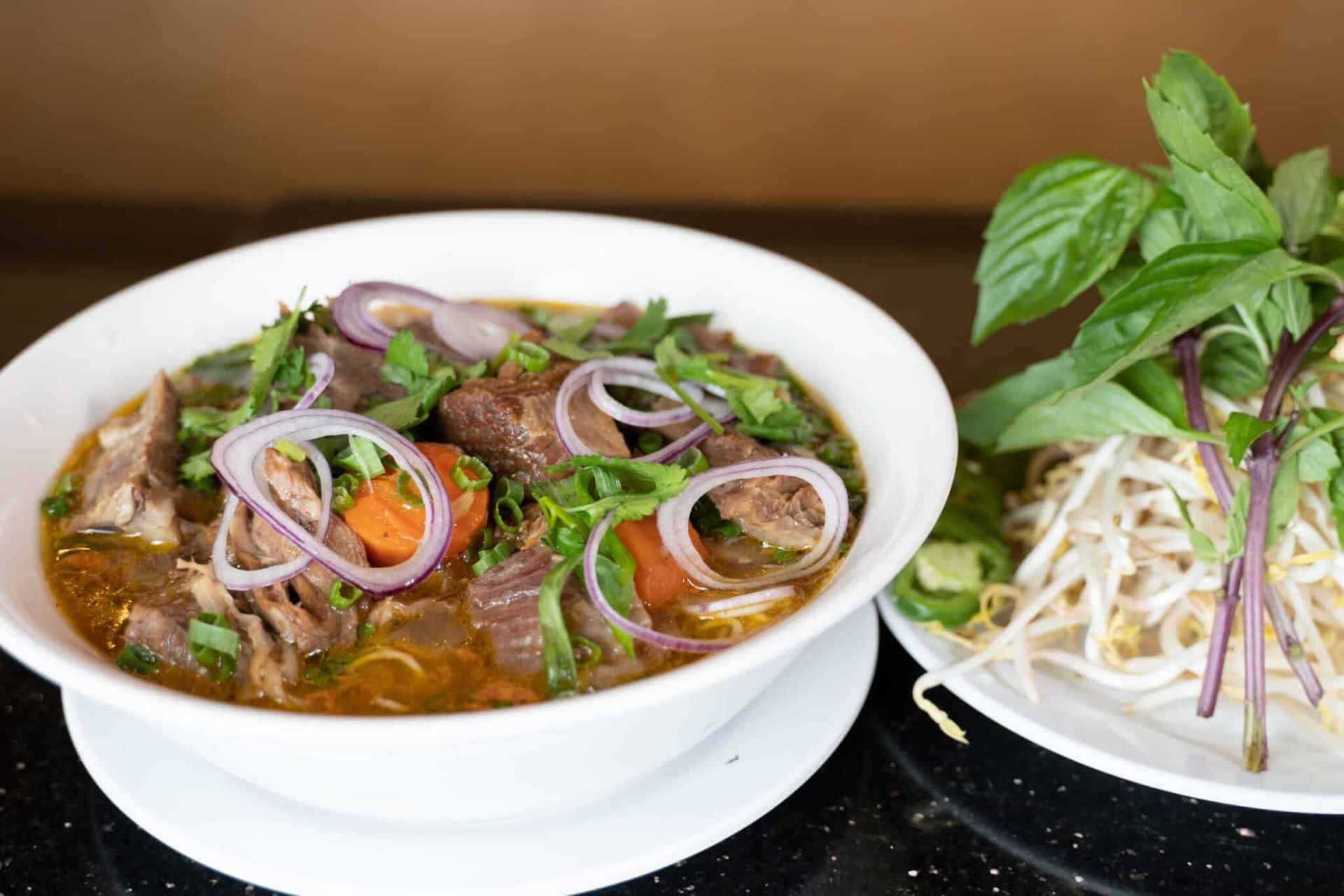 Bowl of beef noodle soup with herbs and onions beside a plate of bean sprouts and basil