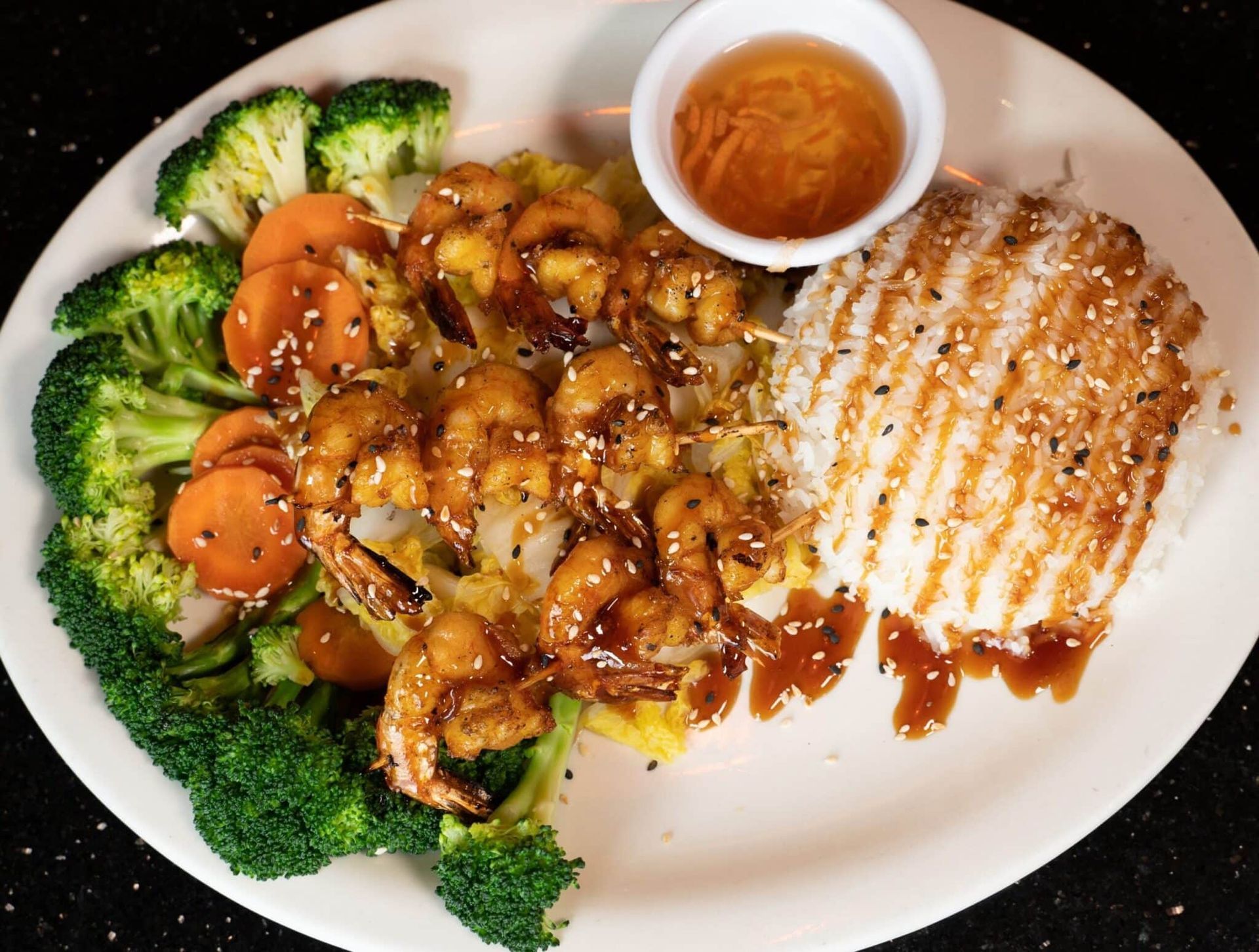Grilled chicken with broccoli and carrots, topped with sauce, on a white plate with a dipping cup