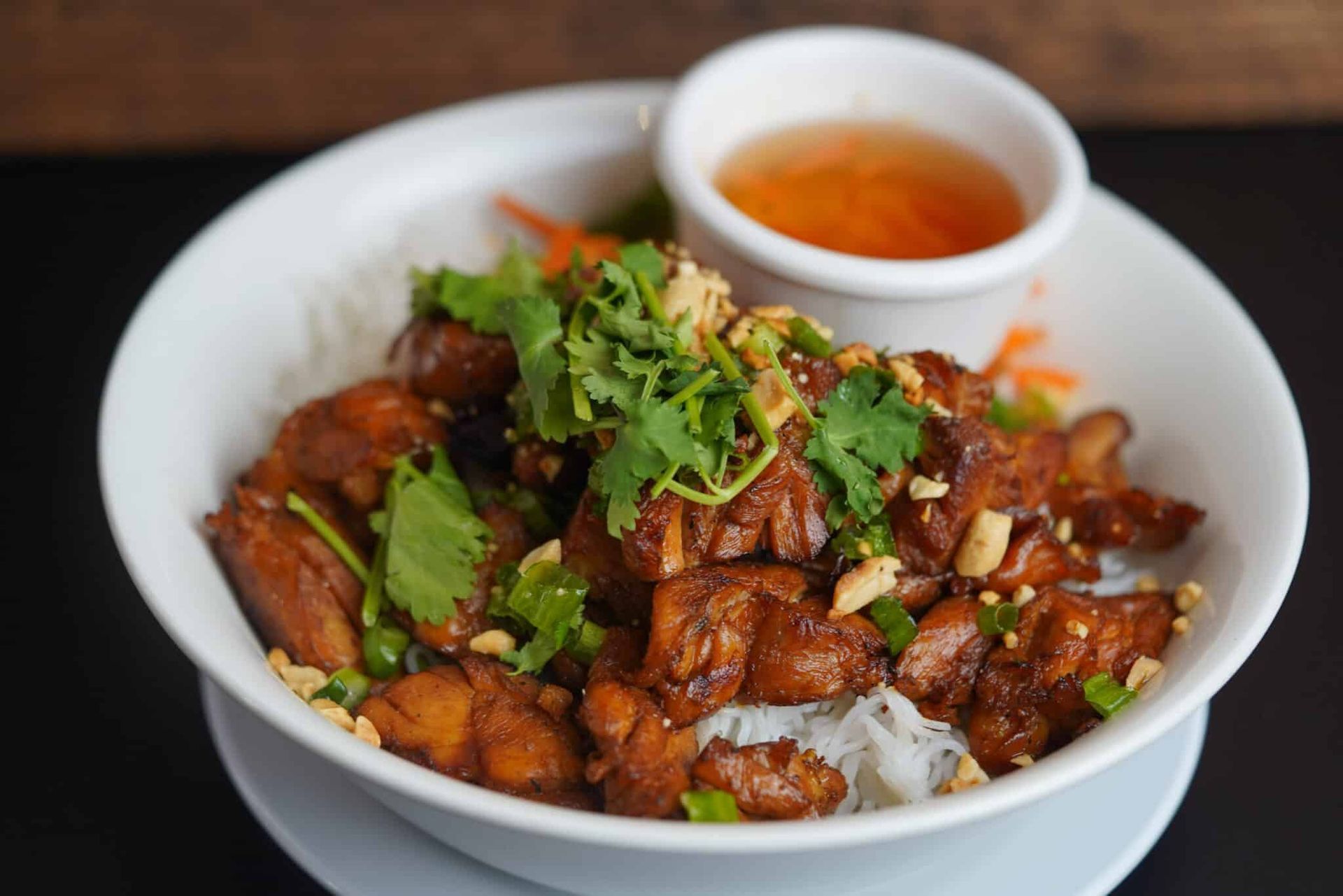 White bowl of saucy browned meat topped with cilantro, with dipping sauce on a dark table