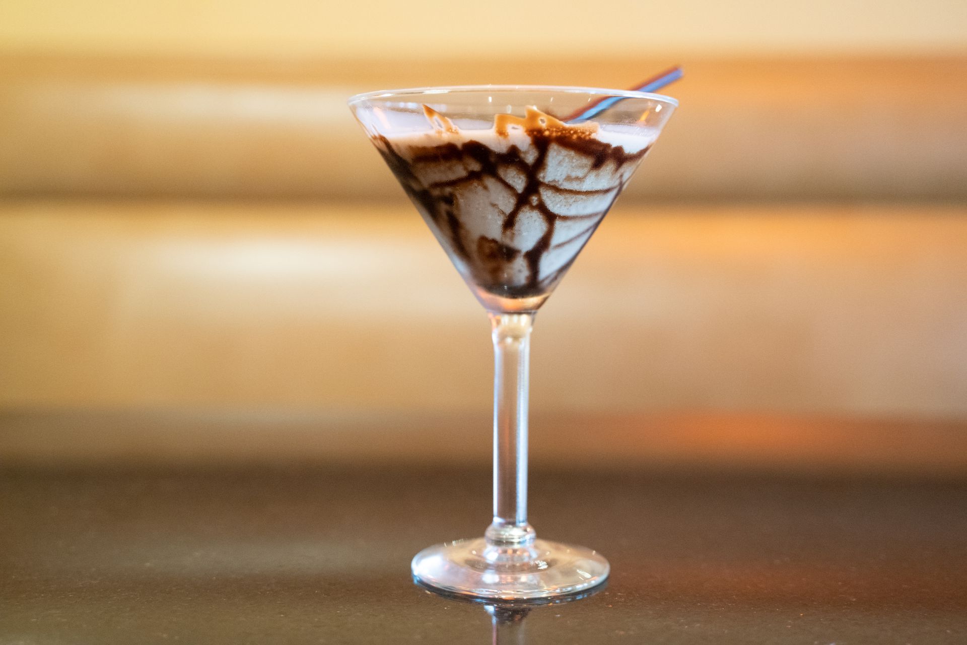 Martini glass with chocolate-drizzled dessert or cocktail on a bar counter, against a warm blurred background