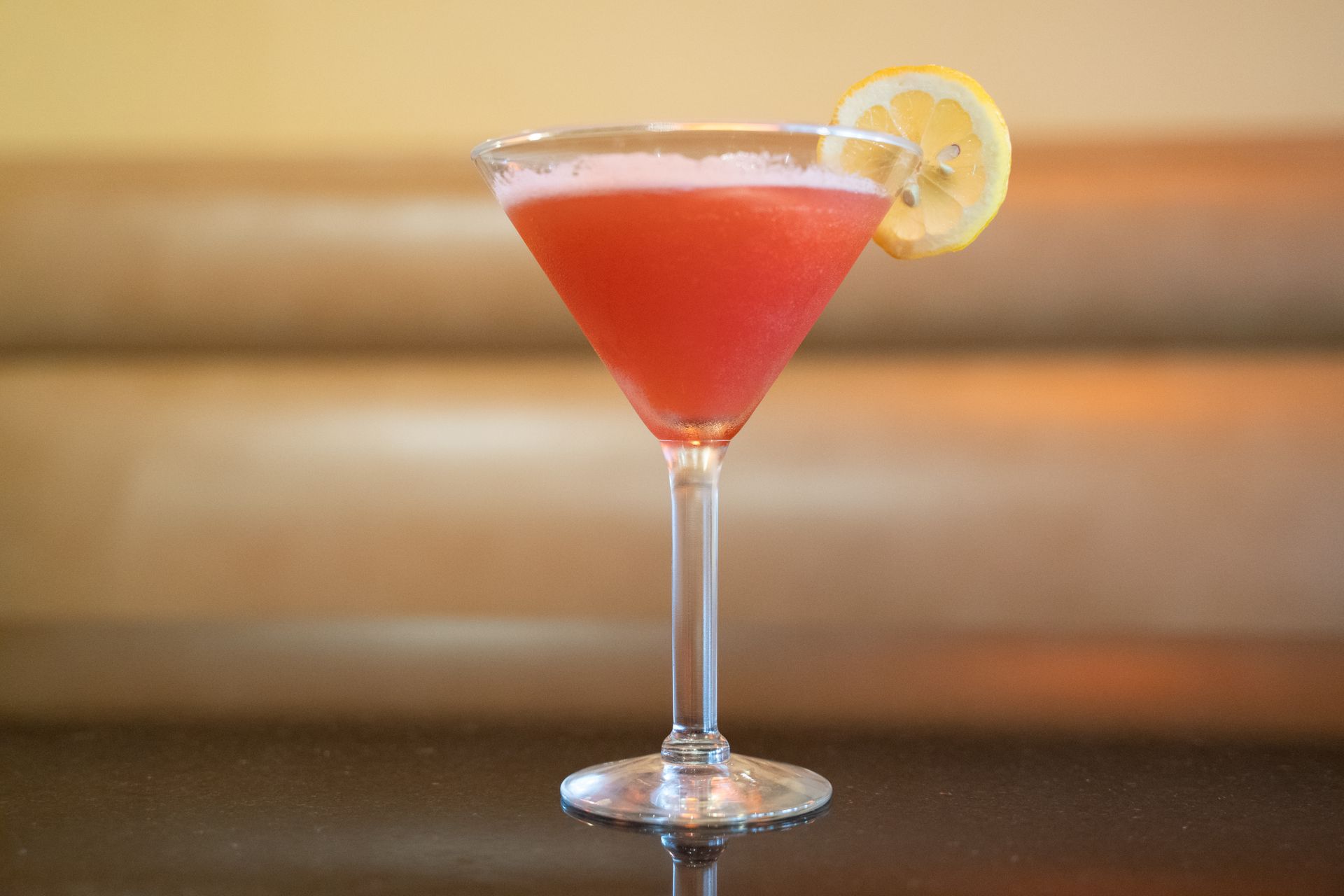 Red cocktail in a martini glass garnished with a lemon slice on a bar counter.